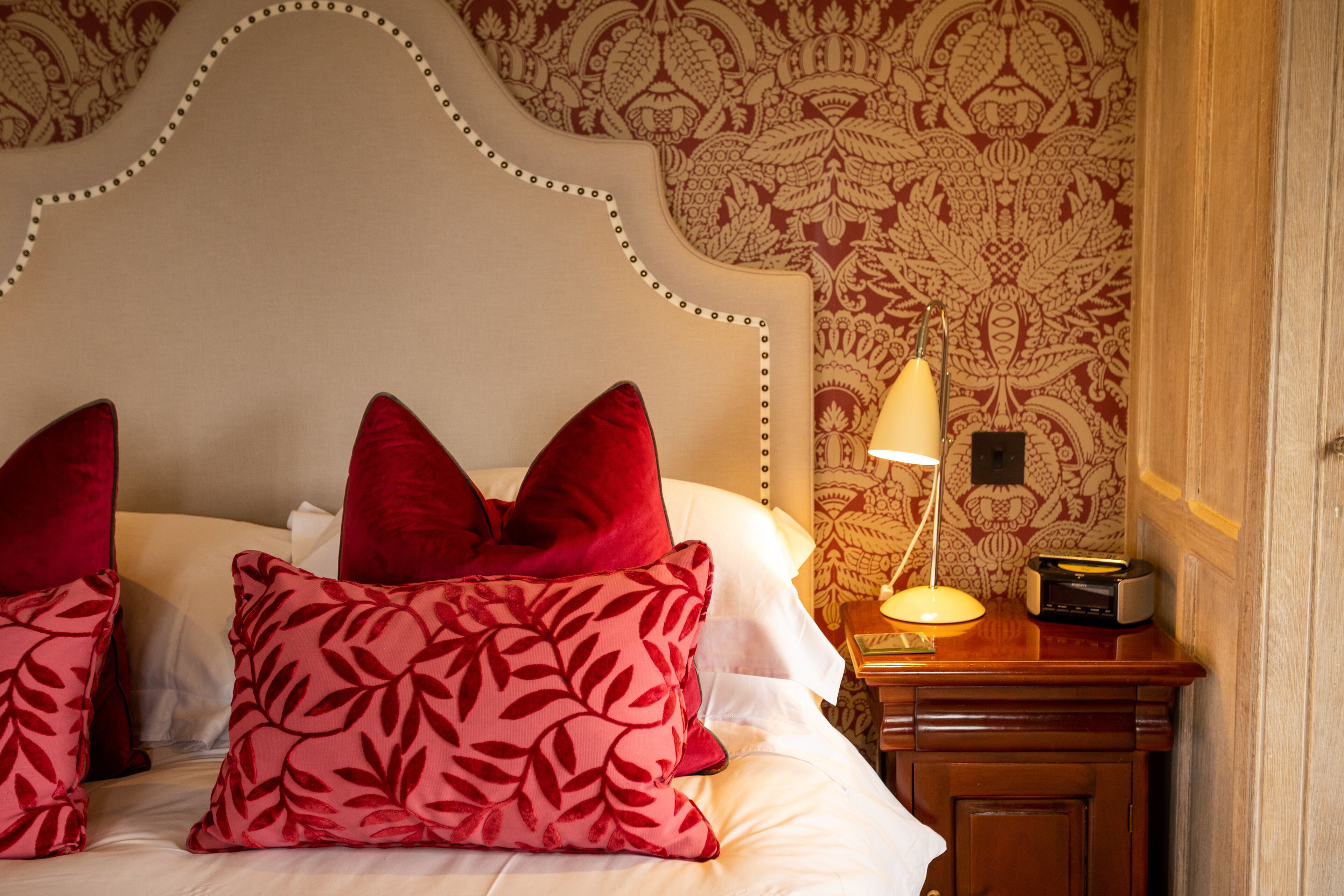 A neatly made bed with red and pink patterned pillows, a classic headboard, and a bedside table with a lamp and radio.