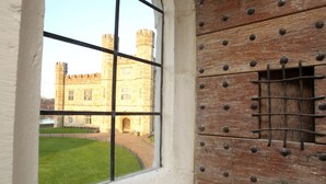 Une fenêtre donnant sur un château, une cour verdoyante et une porte en bois ornée de clous en métal.