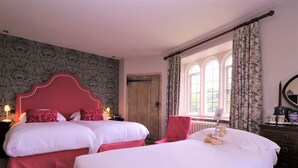 Une chambre d’hôtel avec deux lits, une tête de lit rouge, une porte en bois, une fenêtre avec des rideaux et un ours en peluche sur le lit.