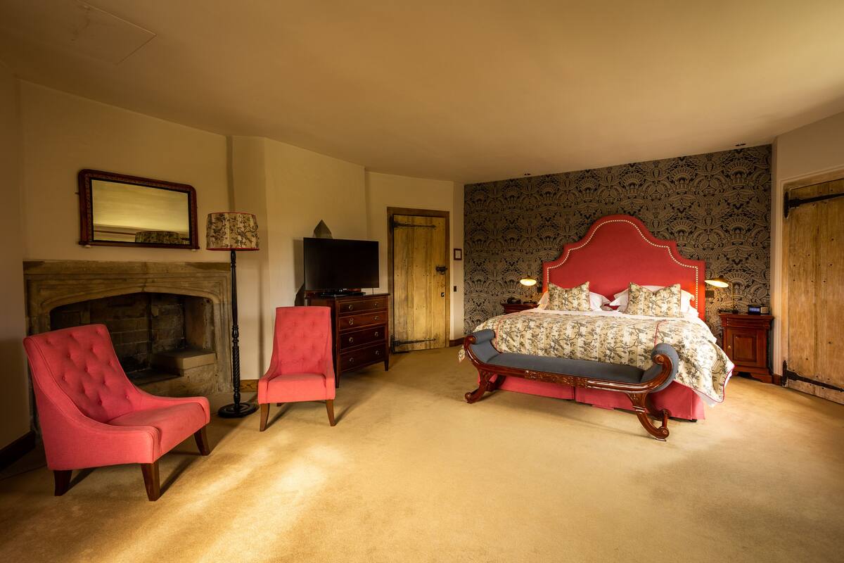 A bedroom with a fireplace, a bed with a red headboard, a red armchair, and a television.