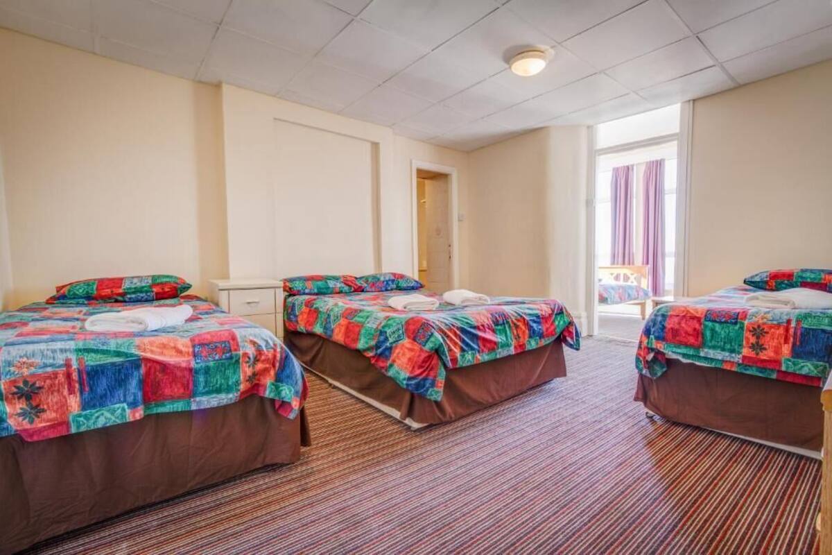 A hotel room with three beds, each with colorful quilts, a small bedside table, and a window with curtains.
