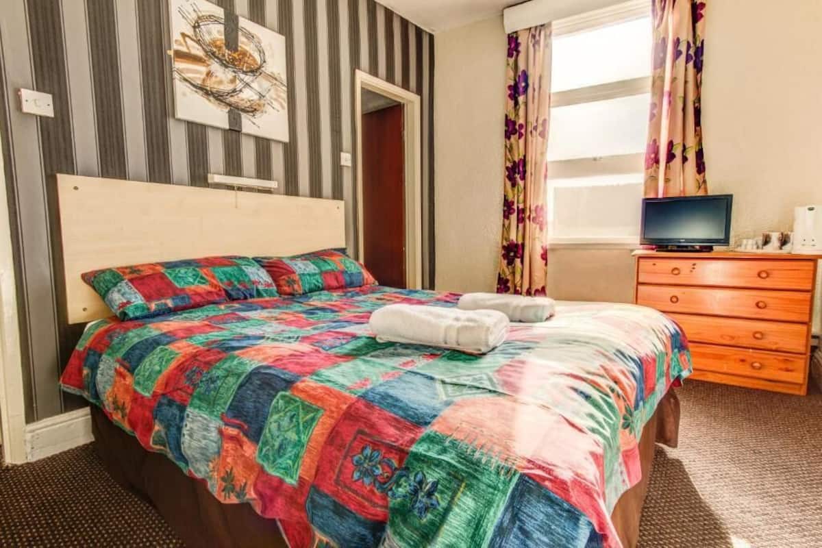 A bedroom with a colorful quilted bedspread, a wooden headboard, a television on a wooden dresser, and a window with floral curtains.
