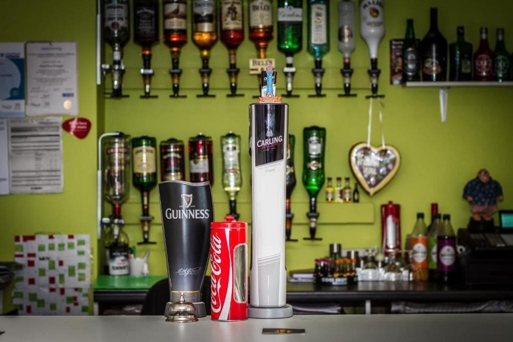 A bar counter with a Guinness beer bottle, a Coca-Cola can, and a Carling beer bottle. Behind the counter, there are various bottles and a heart-shaped decoration on the wall.