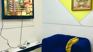 A hotel room with a flat-screen TV mounted on the wall displaying a TV guide, a blue sofa with a yellow pillow, a small table with a flower arrangement, and a framed picture on the wall. - 1 Bed Studio Sleeps 2 - Wifi, Balcony, Parking (Douala)