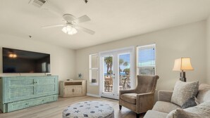 A living room with a flat-screen TV, a ceiling fan, a wooden cabinet, a beige armchair, a round ottoman with nautical prints, and a view of the outdoors through large windows.
