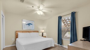 A hotel room with a bed, a ceiling fan, a framed picture, a nightstand with a lamp, a window with curtains, and a TV.