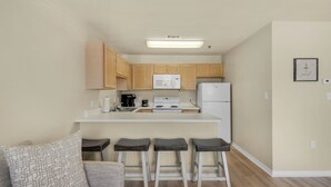 A compact kitchen with wooden cabinets, a microwave, a stove, and a refrigerator. There are three bar stools and a couch with a patterned cushion.