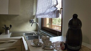 Une cuisine avec un évier, deux tasses posées sur le comptoir, une bouilloire et une fenêtre avec un rideau.