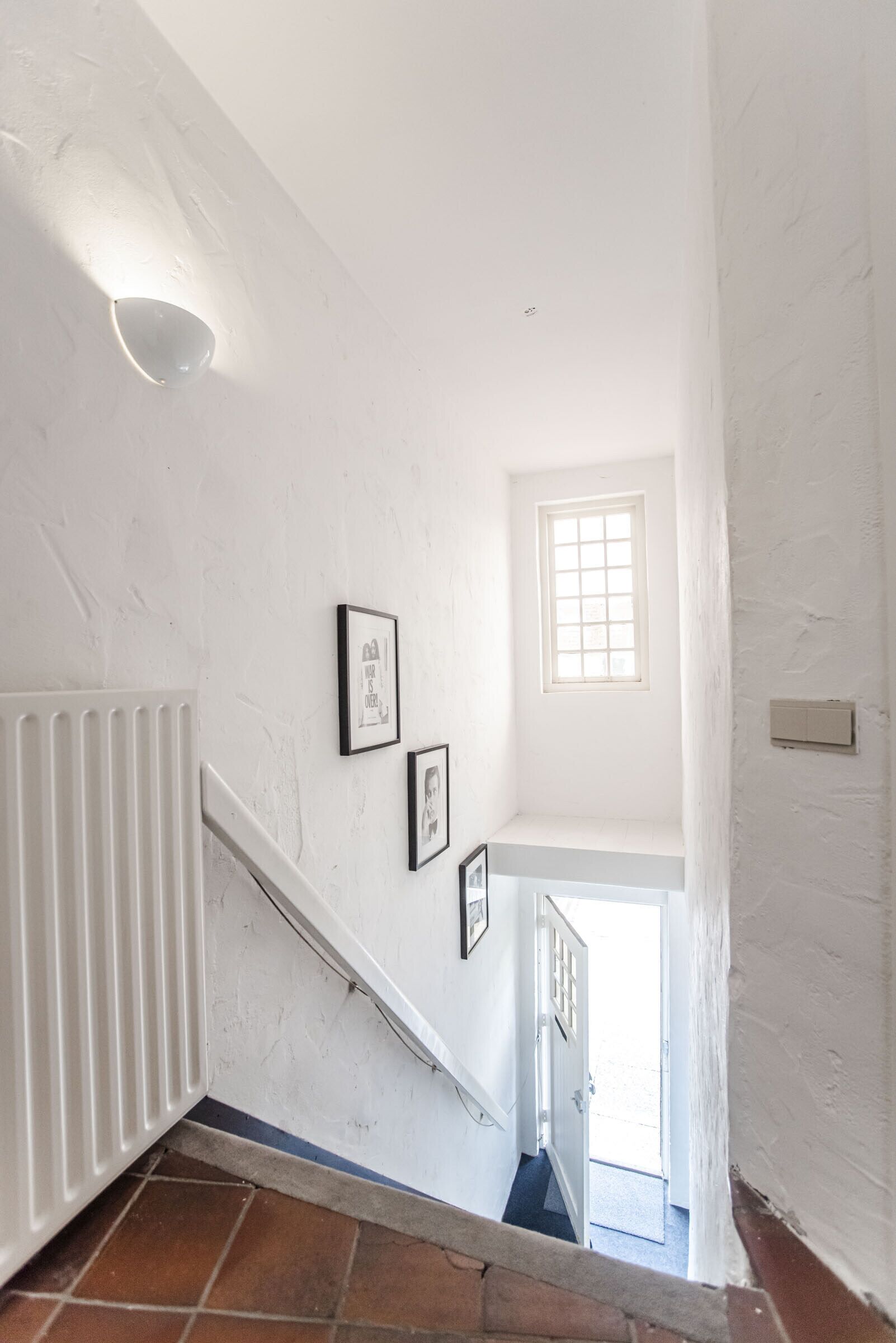 Una escalera blanca con un radiador, una ventana y dos cuadros en la pared.