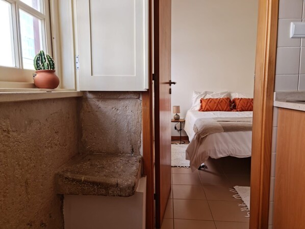 Ein schmaler Flur, der zu einem Schlafzimmer mit Bett, Nachttisch und einer Zimmerpflanze führt.