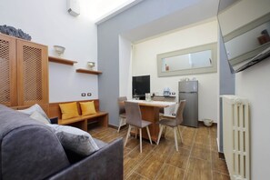 En kompakt opholdsstue med sofa, spisebord, stole, et fjernsyn og et tekøkken med køleskab og mikroovn.
