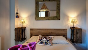 Une chambre à coucher avec une tête de lit en bois, un grand miroir, deux tables de chevet avec des lampes, un lit avec du linge de lit blanc et un sac violet en crochet posé sur le lit.
