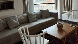 A modern living room with a sofa, a white chair, a small table, and a view of a cityscape through a large window. - Amazing apartment in the best location! (תל אביב-יפו)