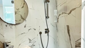 A bathroom with a marble wall, a round mirror, a sink, and a shower with a handheld showerhead. - Amazing apartment in the best location! (תל אביב-יפו)