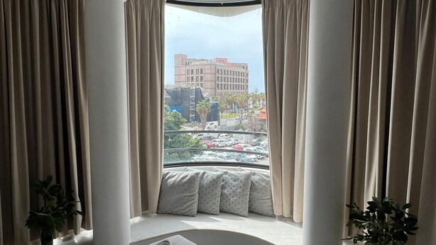 A room with a curved window, beige curtains, a white sofa, and a view of a cityscape. - Amazing apartment in the best location! (תל אביב-יפו)