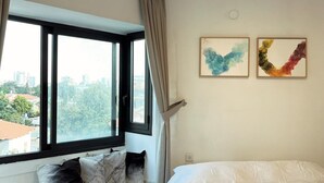 A bedroom with a large bed, a window with curtains, a bench with pillows, and two abstract paintings on the wall. - Amazing apartment in the best location! (תל אביב-יפו)