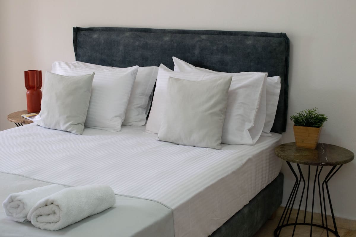 A neatly made bed with white linens and pillows, a small round table with a plant, and a red vase on the side.