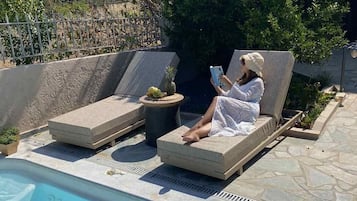 Una persona descansando en una tumbona junto a una piscina, con un libro, rodeada de follaje y un edificio de piedra al fondo.
