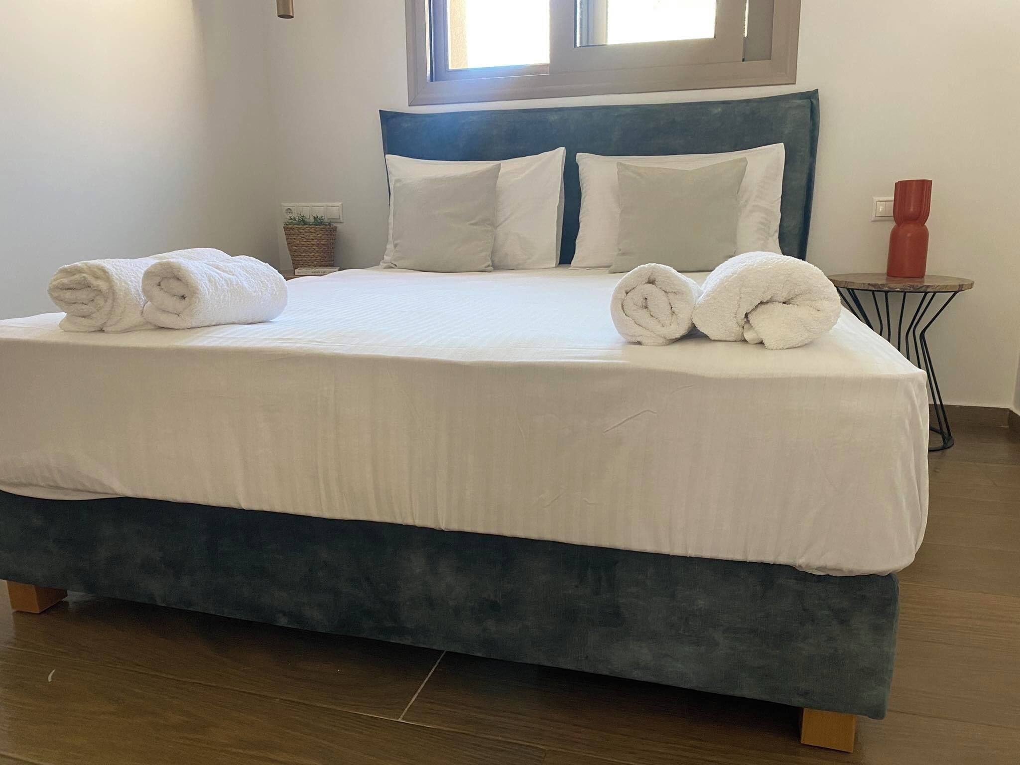 Un lit bien fait, avec du linge de lit blanc et des serviettes pliées, une tête de lit bleue et une petite table de chevet sur laquelle est posé un vase rouge.