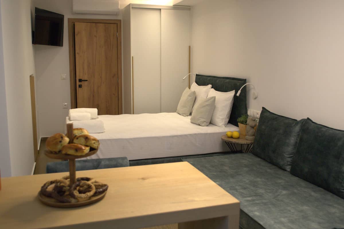 A bedroom with a bed, a bedside table with a tray of pastries, a green sofa, a wooden door, and a TV mounted on the wall.
