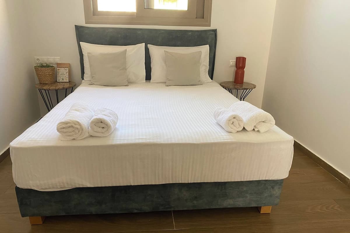 A neatly made bed with white linens and rolled towels, a blue headboard, and a small bedside table with a red vase and a basket.