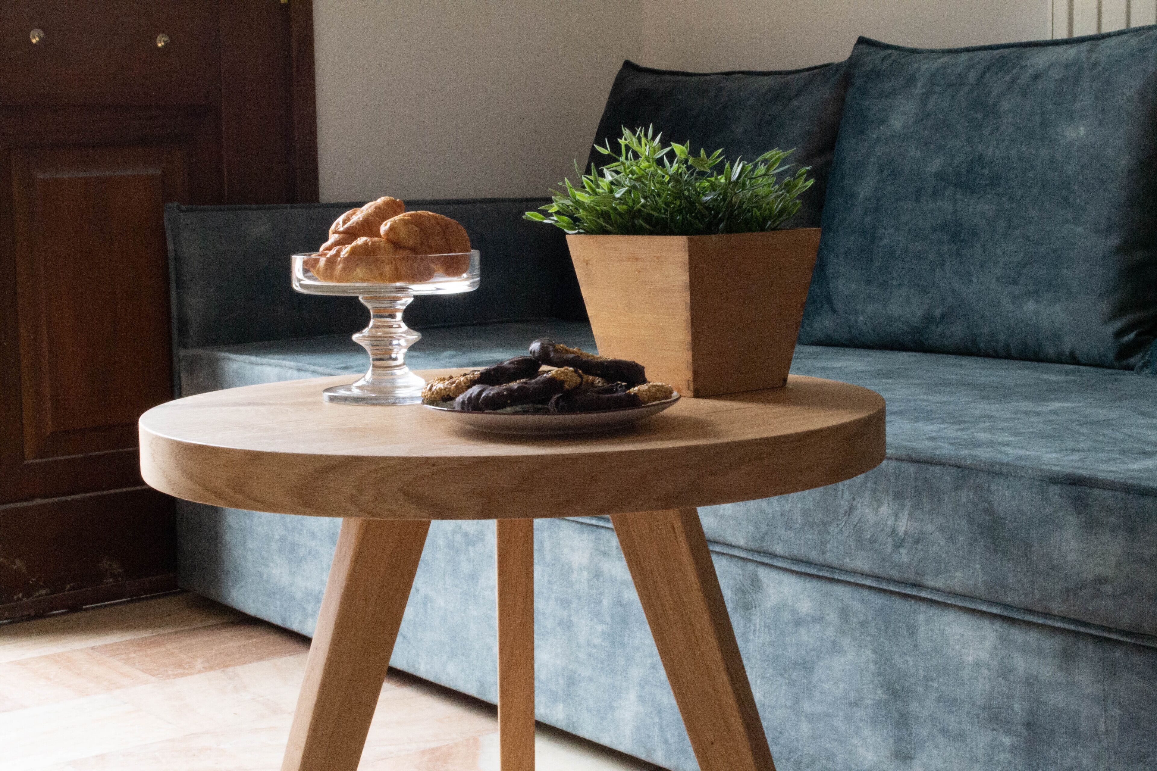 A round wooden table with a glass cake stand and a plate of cookies, a potted plant, and a dark blue sofa in the background.