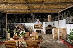 An outdoor patio with a wooden table, chairs, a brick oven, and a cactus plant. - Oniropetra Luxury Suites (Minoa Pediada)