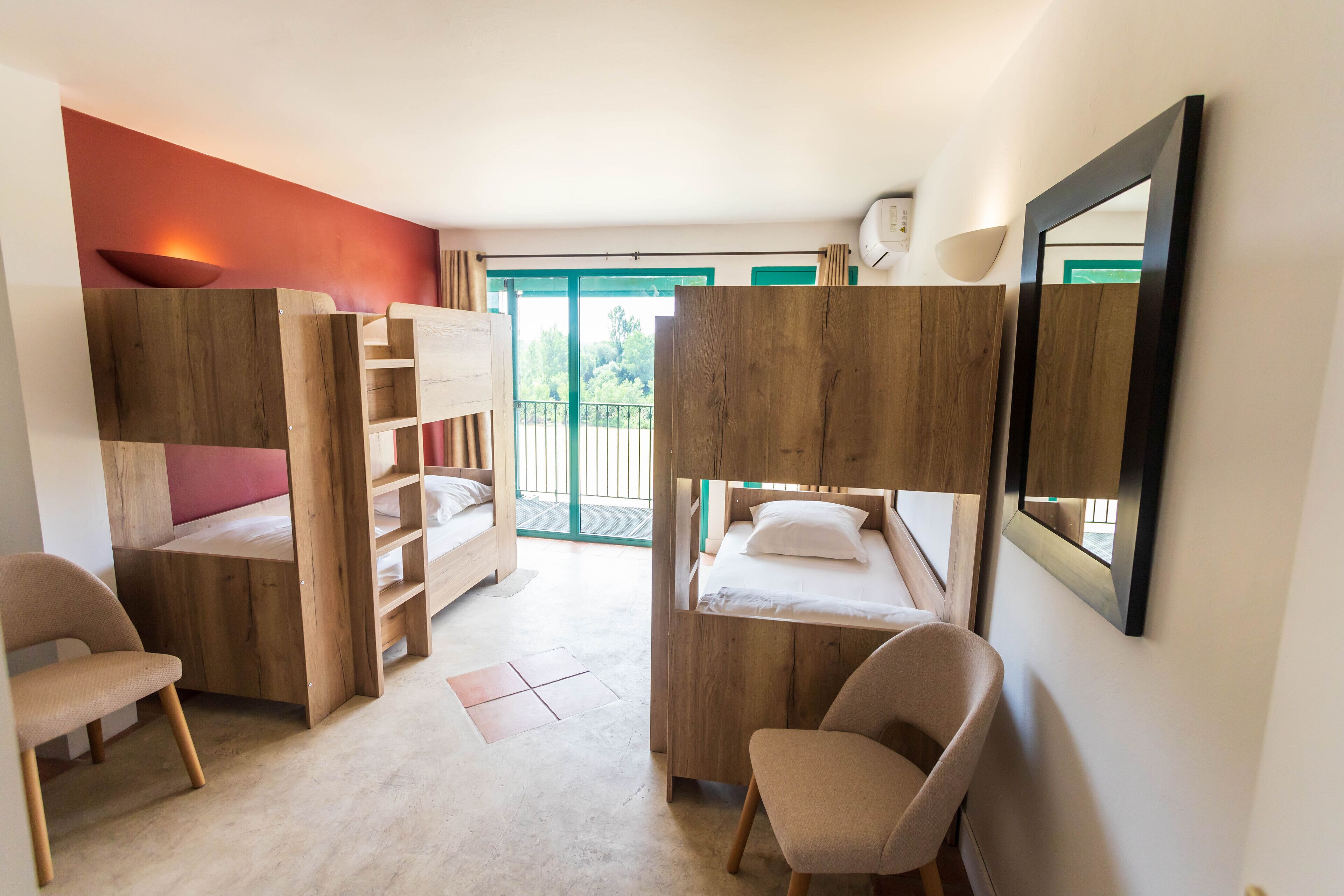 A room with bunk beds, a desk, and a chair. There is a window with a view of greenery outside.