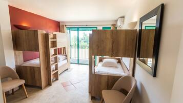 A room with bunk beds, a desk, and a chair. There is a window with a view of greenery outside.