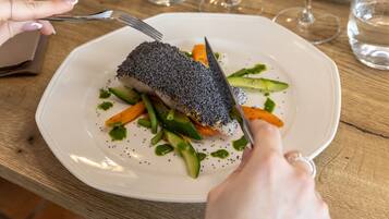 Un piatto bianco con un filetto di pesce cosparso di semi neri, circondato da verdure e salsa, su un tavolo di legno.