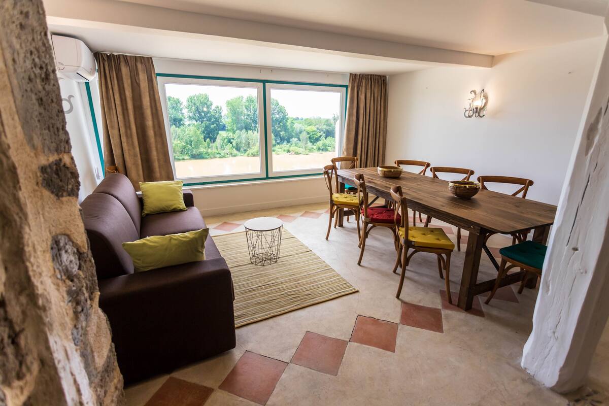 Een eethoek met een houten tafel en stoelen, een bruine bank met groene kussens, en uitzicht op groen door grote ramen.