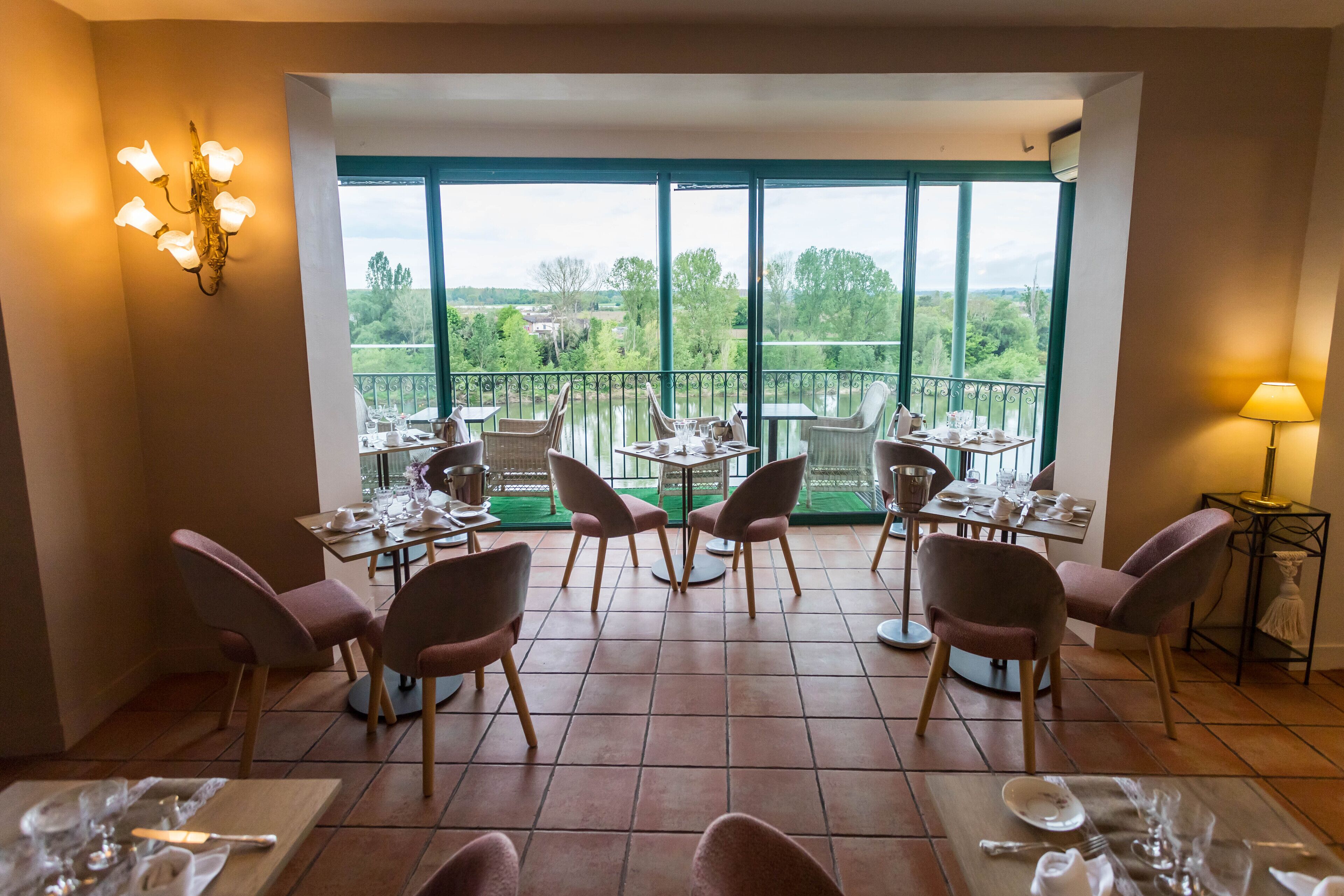 A dining area with tables and chairs, large windows with a view of greenery, and a chandelier.