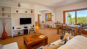 A spacious living area with a flat-screen TV, wooden furniture, and a dining area with a large window. - SES SAVINES A charming, peaceful house with a pool and BBQ, 2km from the beach (Illes Balears)