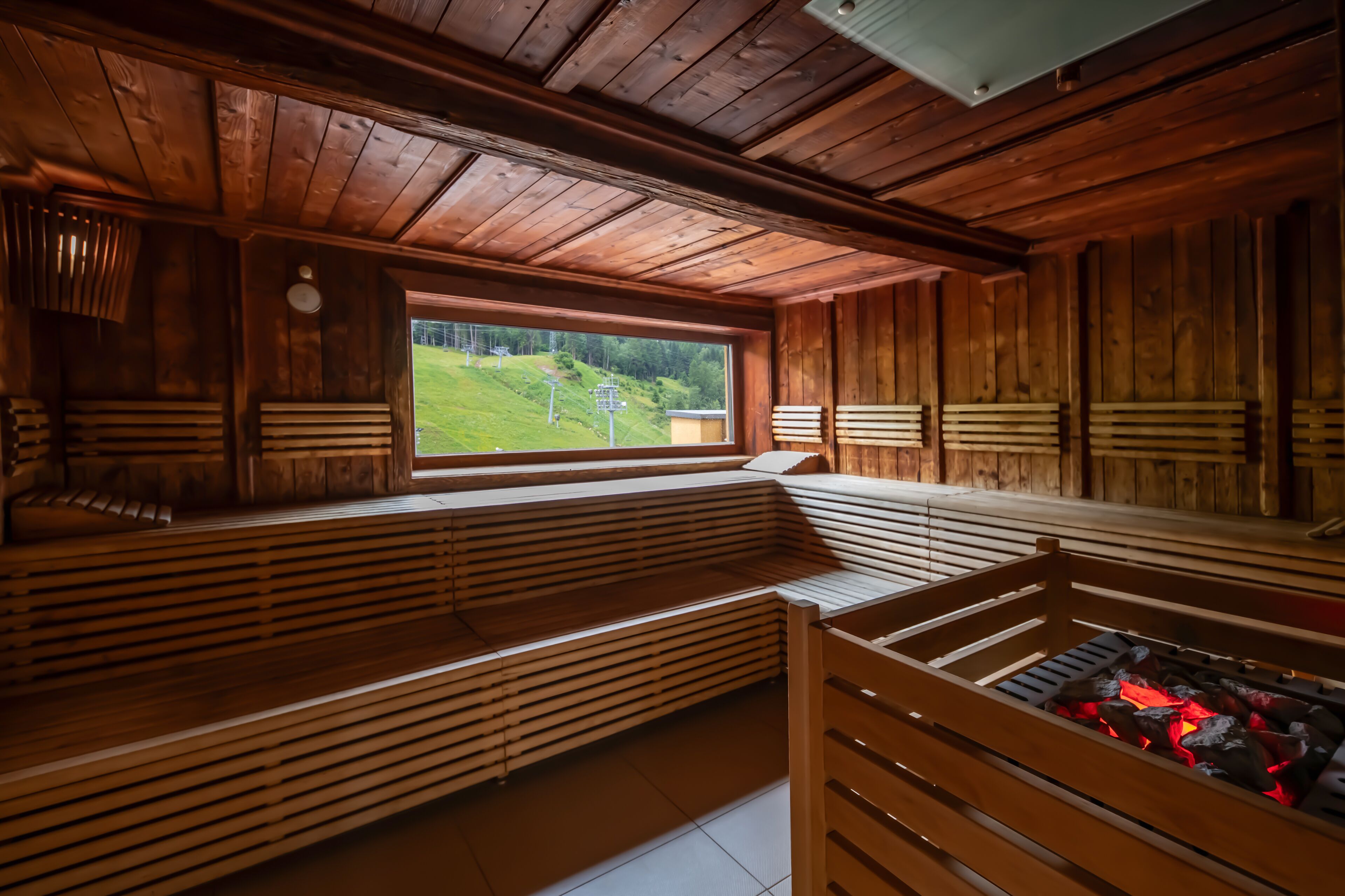 A wooden cabin with a large window offering a view of a green landscape. Inside, there are wooden benches and a fireplace with logs.