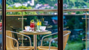Un balcón con sillas de mimbre y una mesa dispuesta para dos, con vistas a un paisaje de vegetación y edificios.