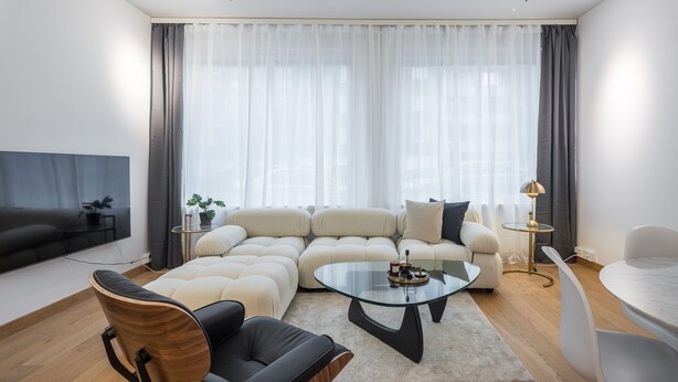 A modern living room with a sectional sofa, a glass coffee table, a black leather armchair, a small side table with a lamp, and a flat-screen TV mounted on the wall. - Nordic townhouse with own sauna at the city center of Helsinki (Helsinki)