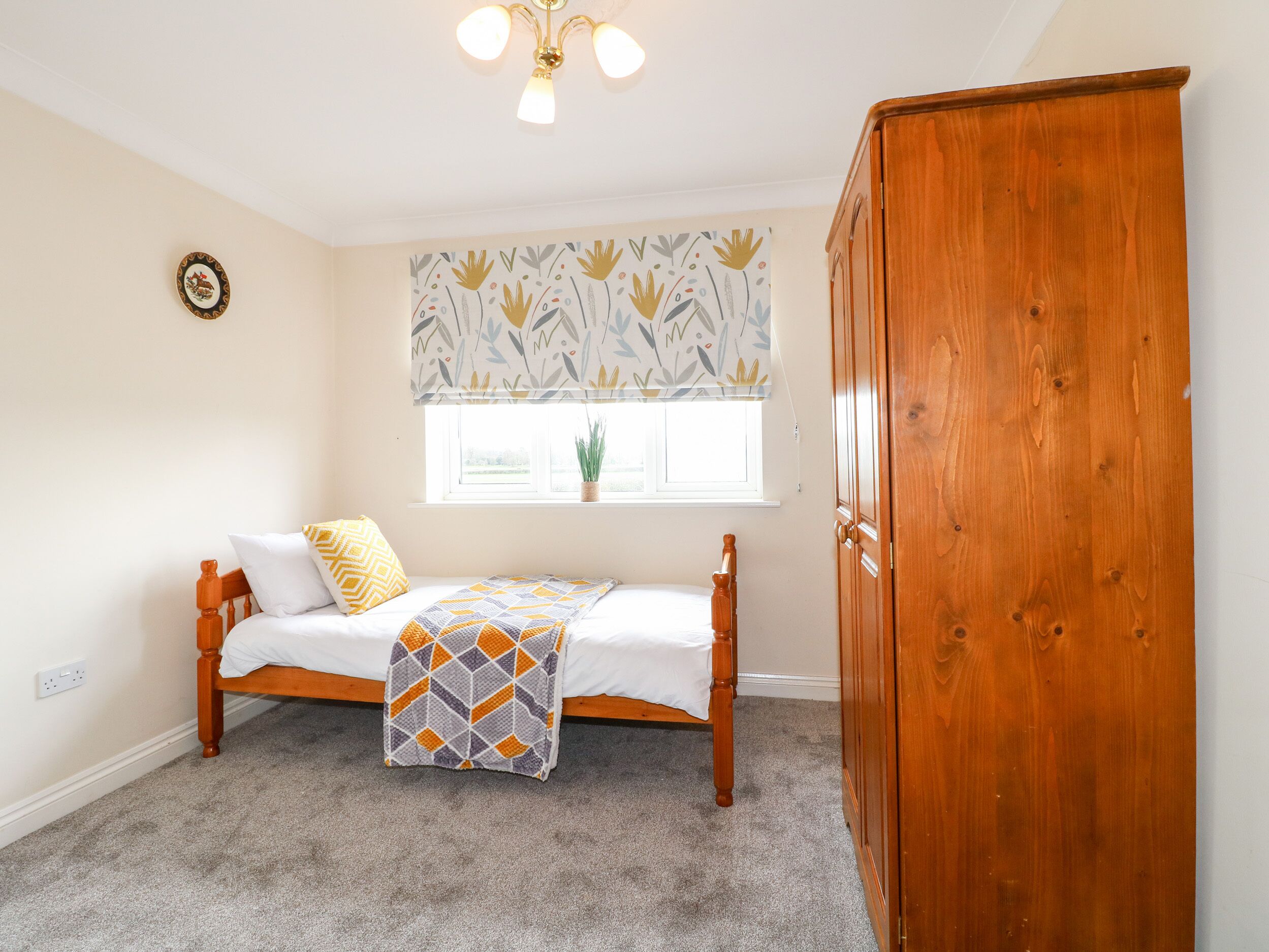 A room with a bed, a wooden wardrobe, a window with a plant, and a ceiling light.