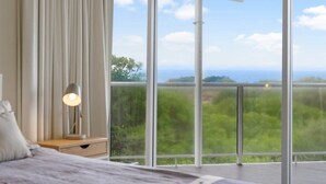 A bedroom with a large window, a bedside table with a lamp, and a view of greenery and a distant landscape.