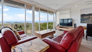 A modern living room with a large TV, leather sofas, a wooden coffee table, and expansive windows offering a view of the landscape.
