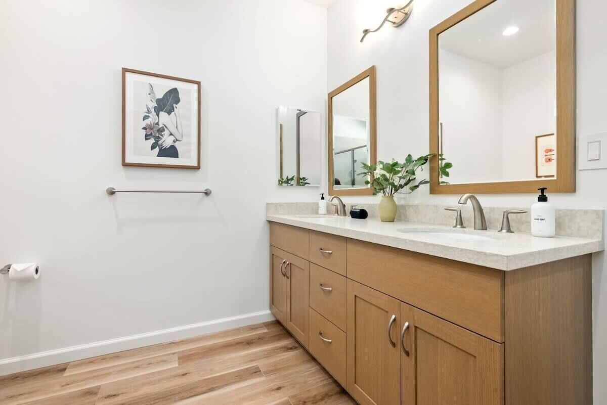 A modern bathroom with a wooden vanity, two mirrors, a framed artwork, and a toilet paper holder.