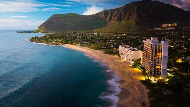 A coastal hotel with a beachfront view, a mountain in the background, and a well-lit building.
