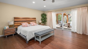 A bedroom with a wooden headboard, a bench, a nightstand, a lamp, and a large window with curtains. - Peaceful Retreat near Lake Travis & Volente (Volente)