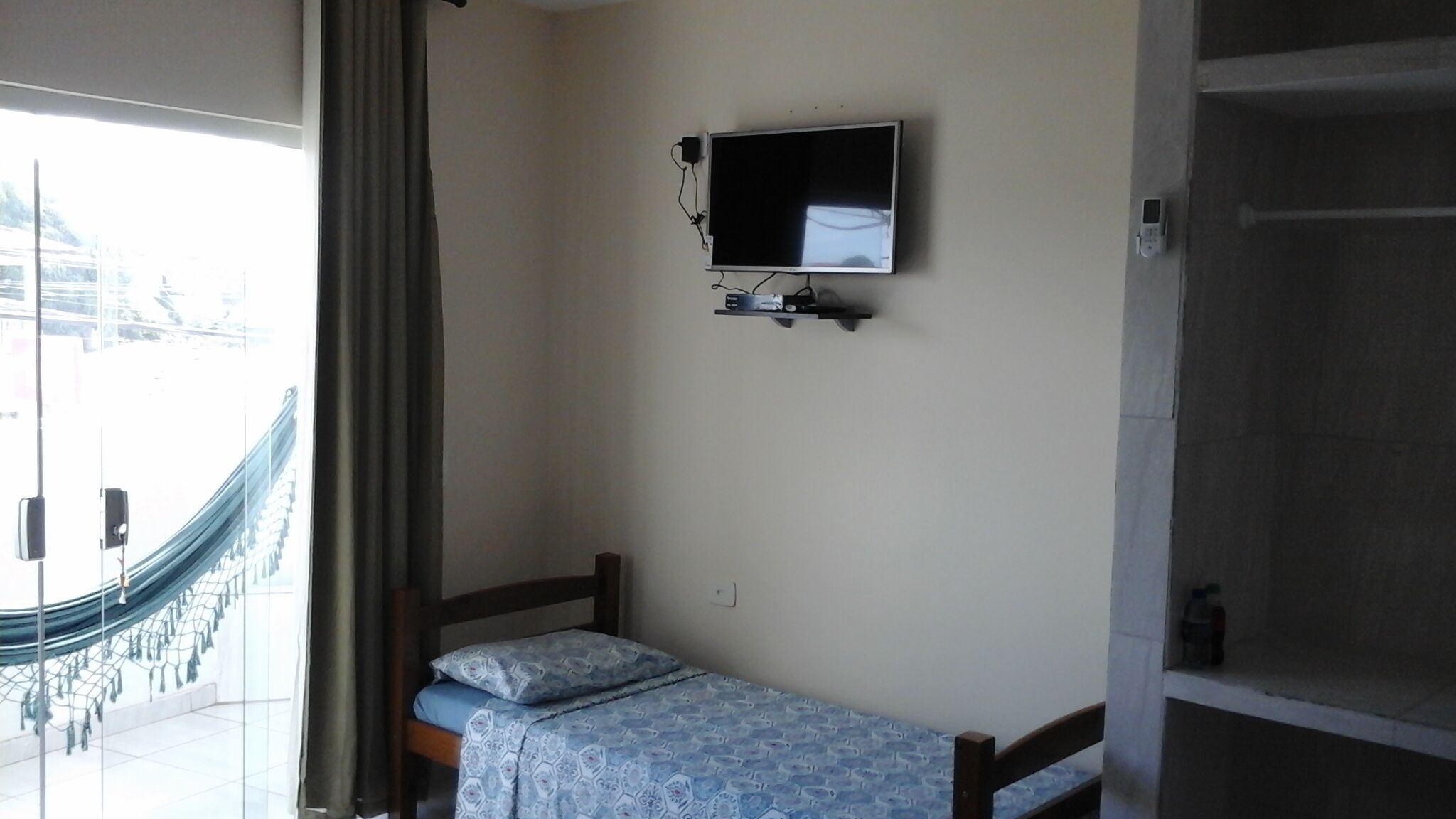 A small room with a single bed, a television mounted on the wall, and a hammock outside the window.