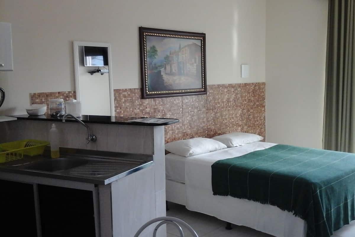 A hotel room with a bed, a small kitchenette, a framed picture on the wall, and a window with curtains.