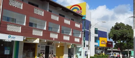 Un immeuble à plusieurs étages abritant une pharmacie au rez-de-chaussée, un restaurant avec une enseigne rouge et jaune au-dessus, et divers commerces, dont une pharmacie et un magasin de téléviseurs à écran plat.