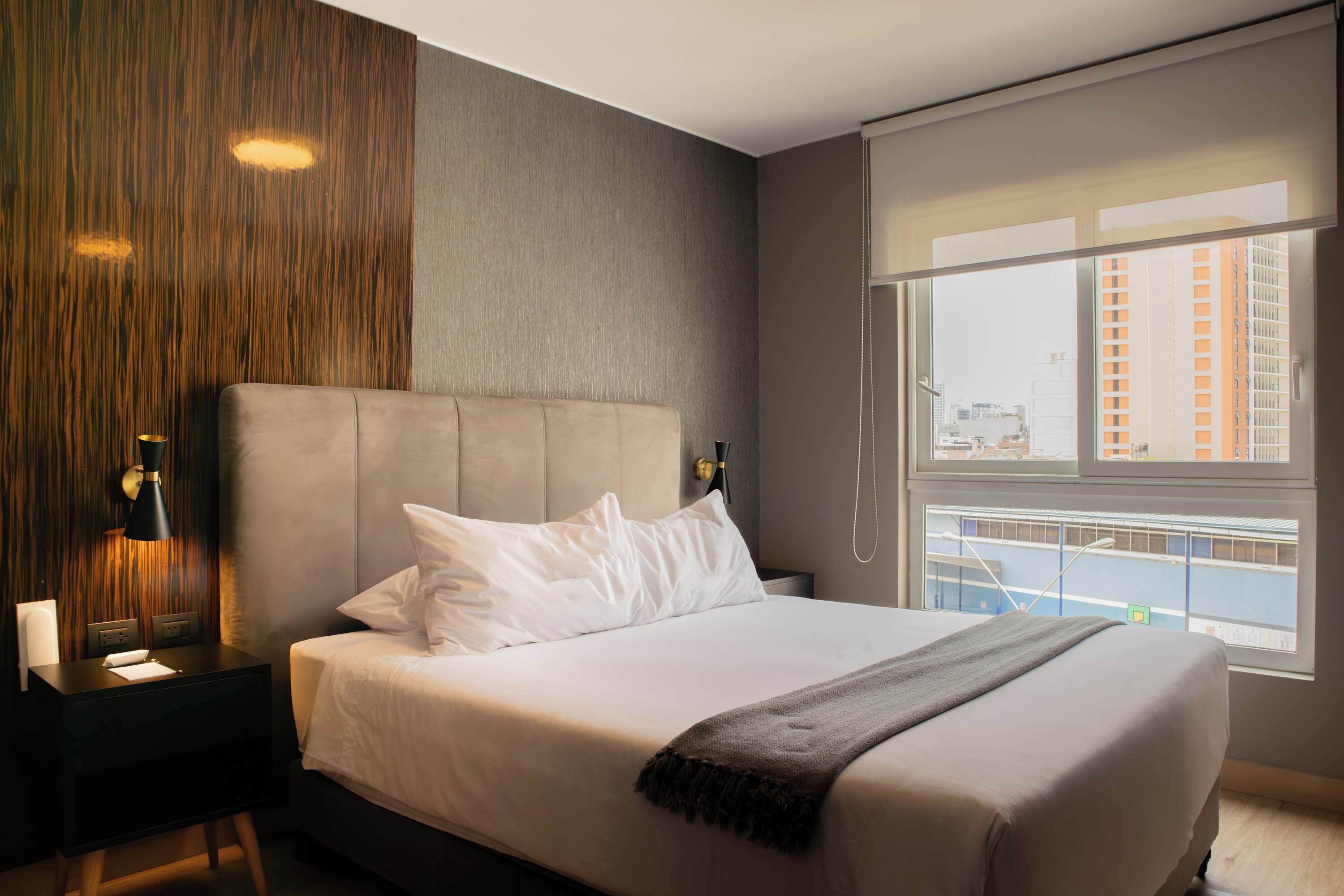 Une chambre d’hôtel moderne avec un grand lit, une table de chevet, une lampe et une vue sur la ville grâce à une grande fenêtre.