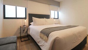 Un dormitorio con una cama grande, dos mesitas de noche con lámparas, un sofá gris y una ventana con persianas.