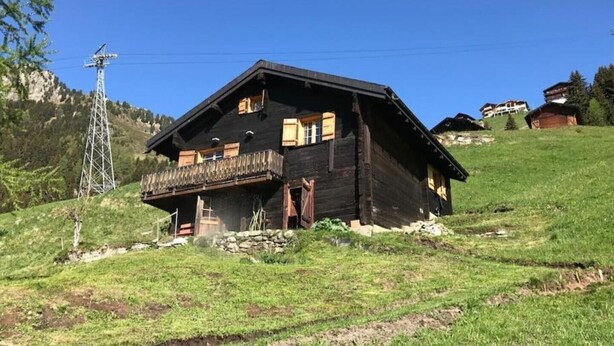 Un chalet in legno con un balcone, situato su un pendio erboso sotto un cielo azzurro.