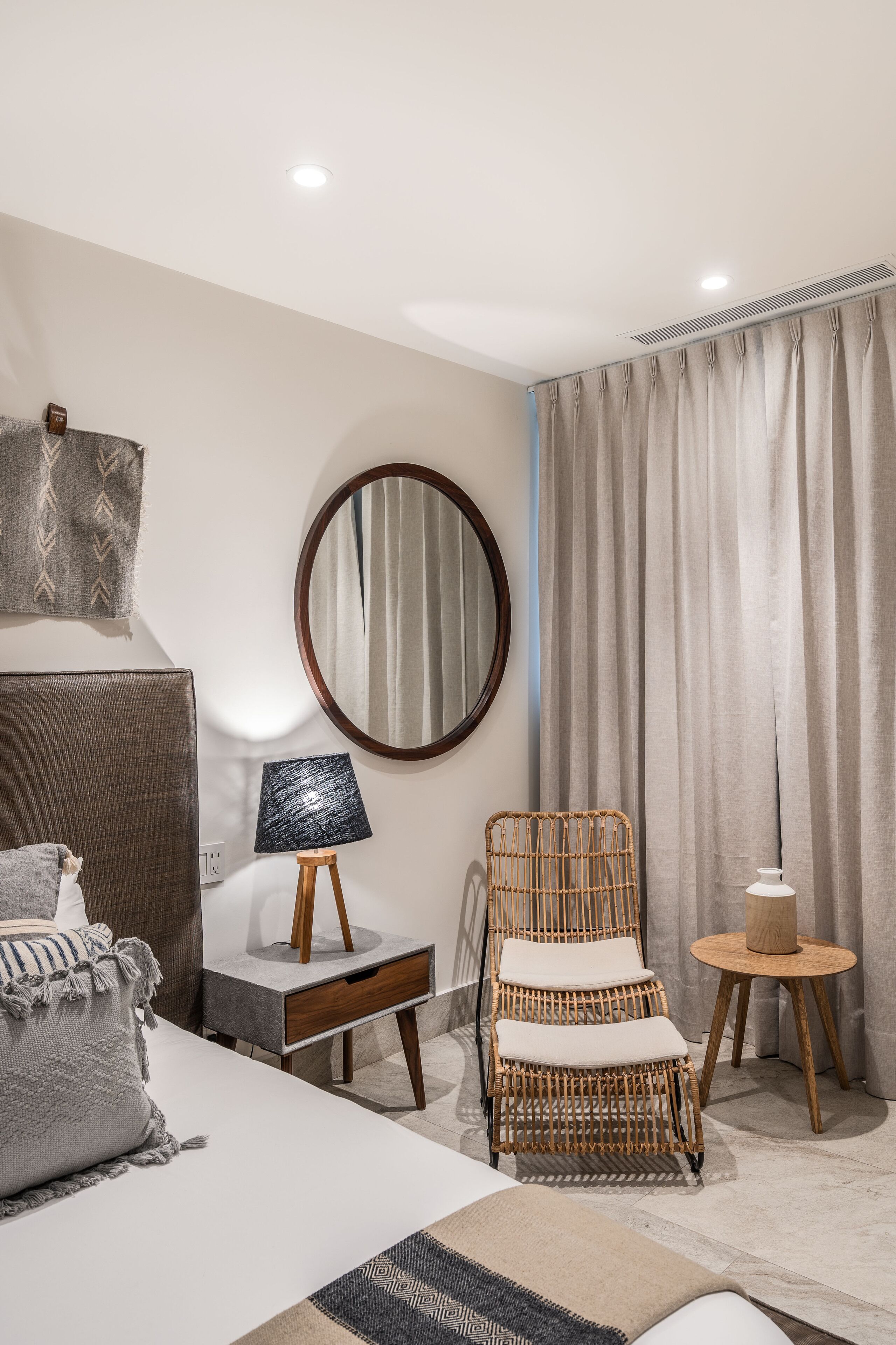 A modern hotel room with a bed, a wicker chair, a small table with a lamp, a mirror, and a large window with curtains.