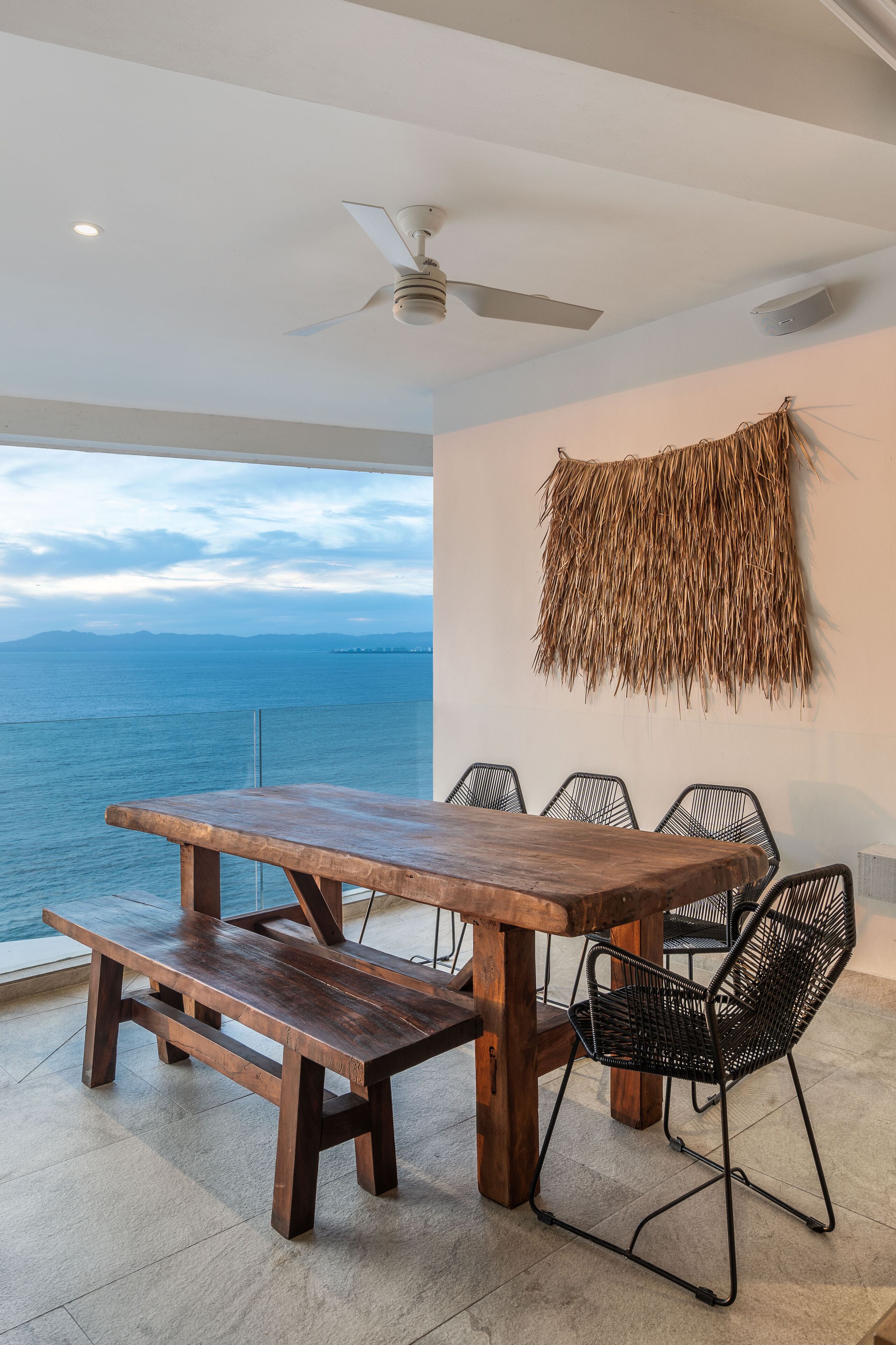 Une table à manger en bois, accompagnée de bancs, de chaises en fil de fer noir et d’un ventilateur de plafond, dans une pièce dotée d’une grande fenêtre donnant sur la mer.